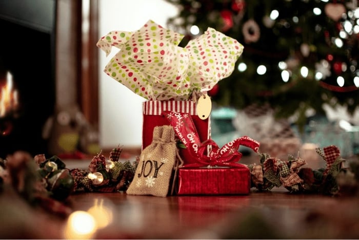 Red gift bags, one with "JOY" on the front, in front of a Christmas tree.