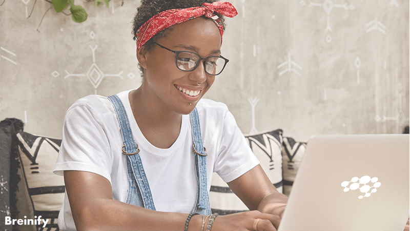 smiling black woman with black glasses, red bandanna in their hair, white tee, and jean overalls working on a laptop