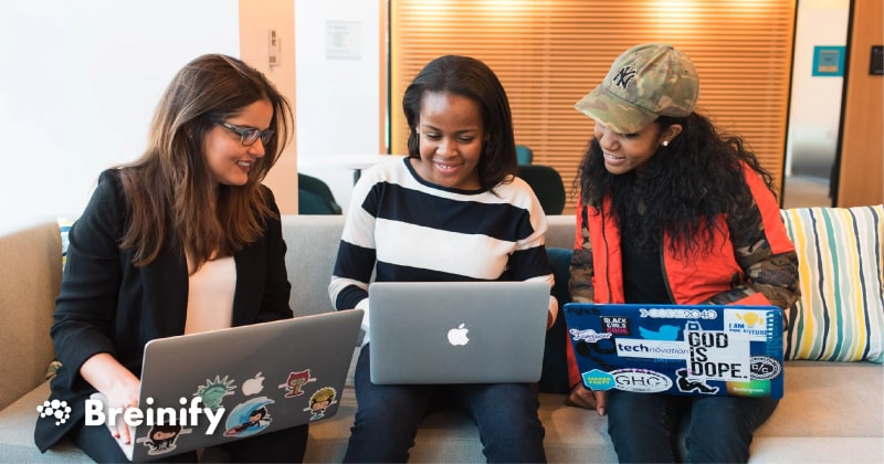 three women sit together on a couch looking at laptops and smiling