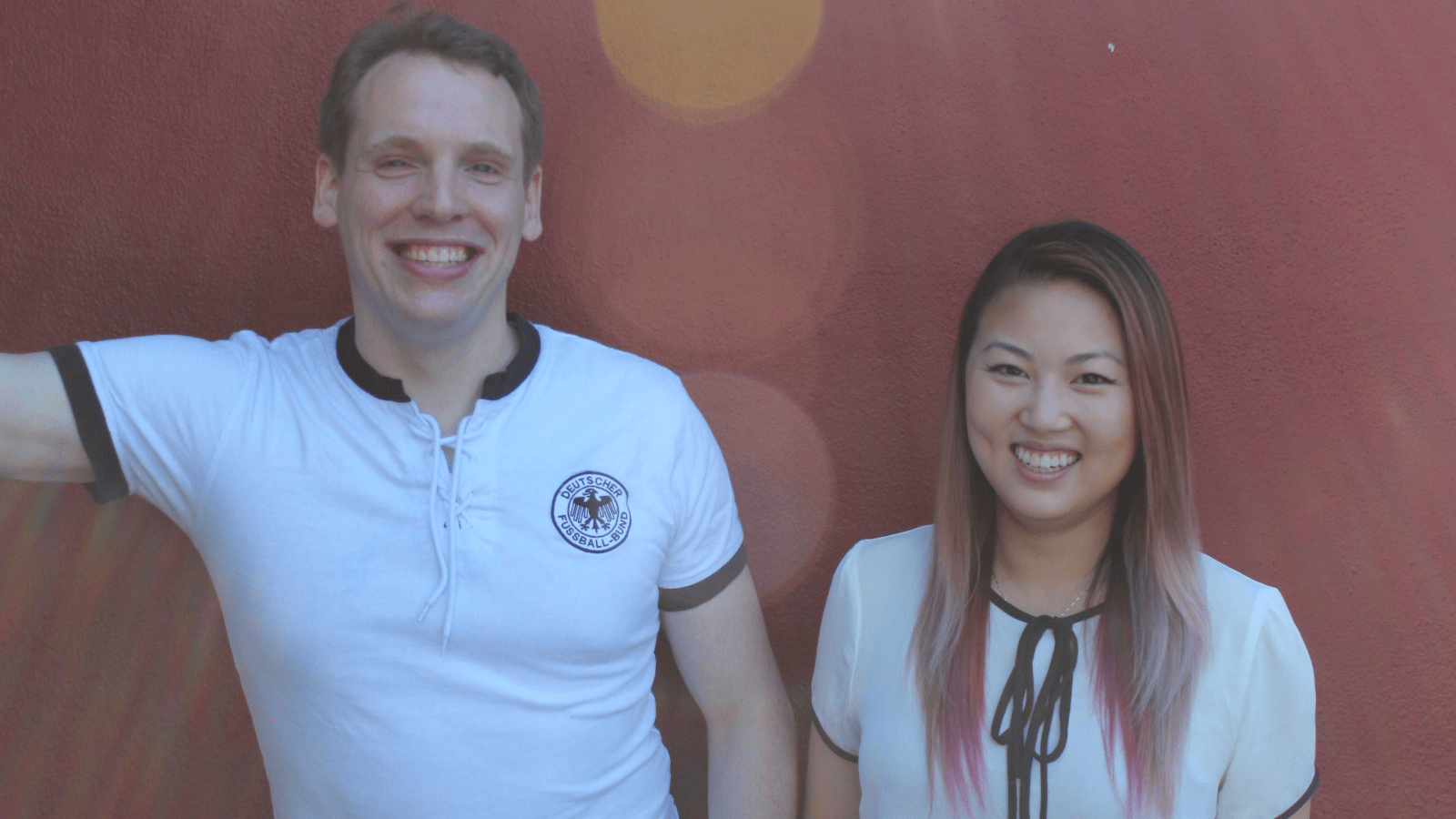 Philipp and Diane wearing white colored tops, smiling in front of a dark maroon wall